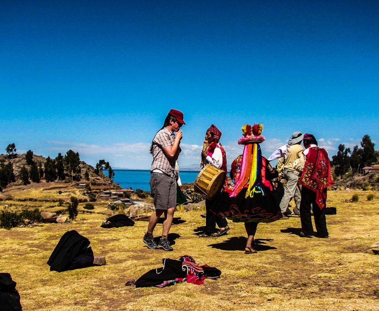 A group of people in traditional Andean clothing perform a cultural dance outdoors near Lake Titicaca, with one man playing a drum and others in colorful attire, under a clear blue sky.