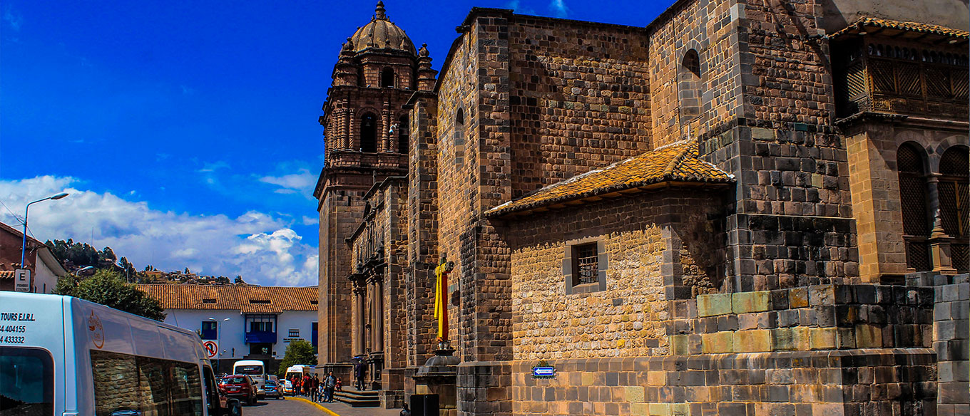 Street view of a stone colonial church in Cusco, with a clear blue sky and vehicles parked nearby.