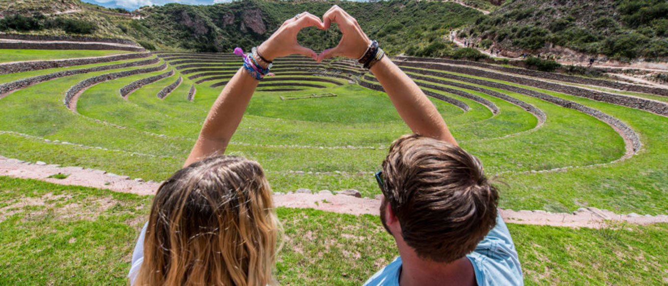 A couple sits facing the circular terraces of Moray, Peru, forming a heart shape with their hands over the green landscape.