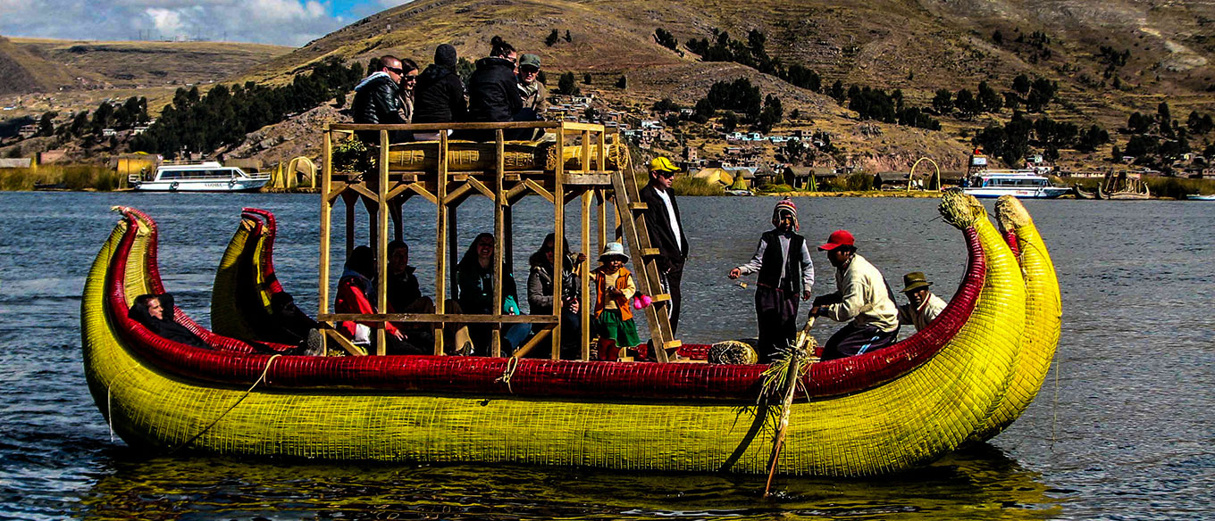 A group of people on a traditional reed boat on Lake Titicaca, with a raised platform and brightly colored yellow and red design, surrounded by hills and small houses in the background.