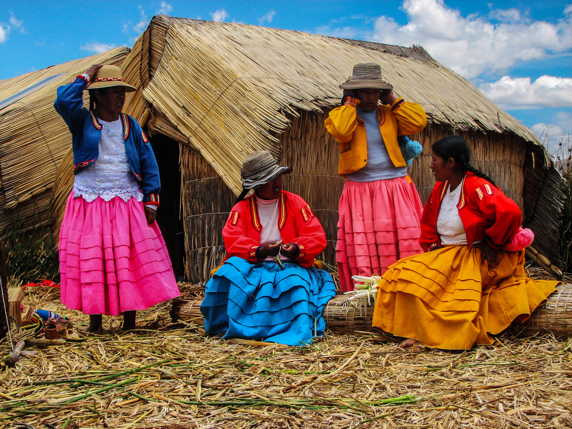 Four women in traditional colorful clothing sitting and standing in front of reed huts on the Uros Islands, Lake Titicaca.