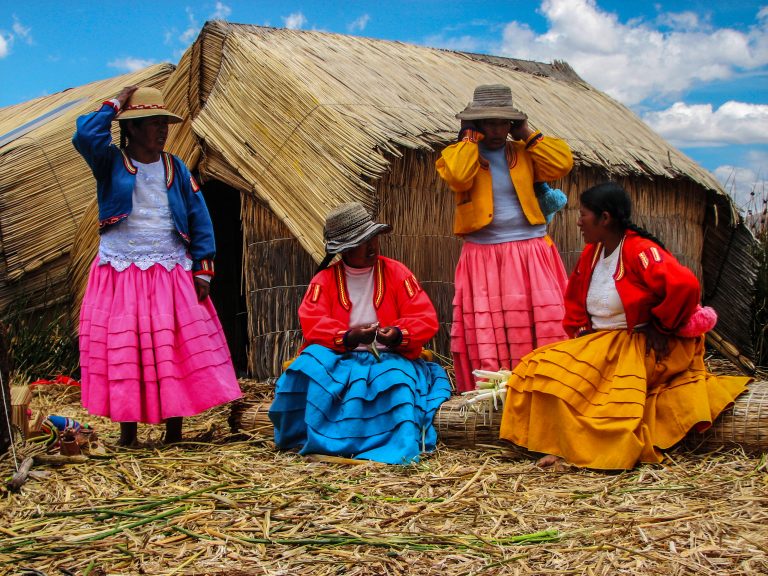 Four women in colorful traditional skirts and hats standing and sitting outside reed huts on Lake Titicaca.