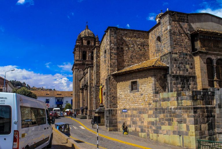 A street view of a stone colonial church in Cusco, Peru, with a tall bell tower, surrounded by parked vehicles and people walking under a bright blue sky with scattered clouds.